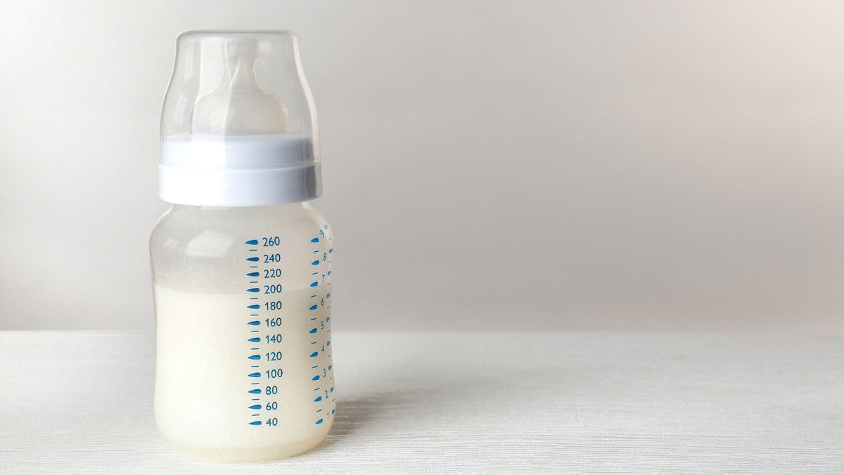 A glass baby bottle with a white cap and teat, part filled, on a plain background