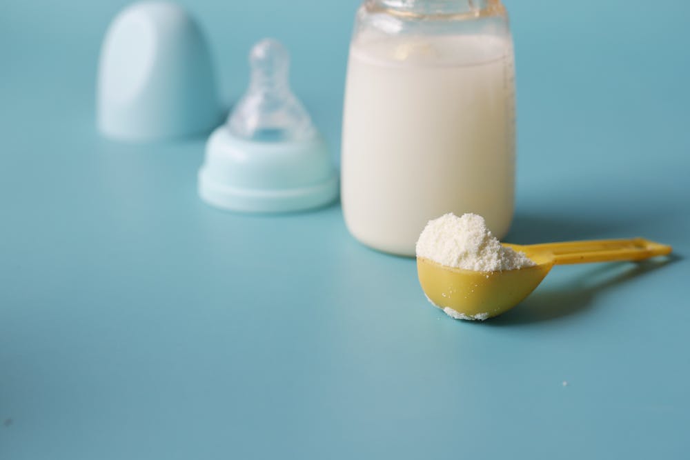 A baby bottle on a pale blue background with its cap and teat beside it and a scoop of formula in front